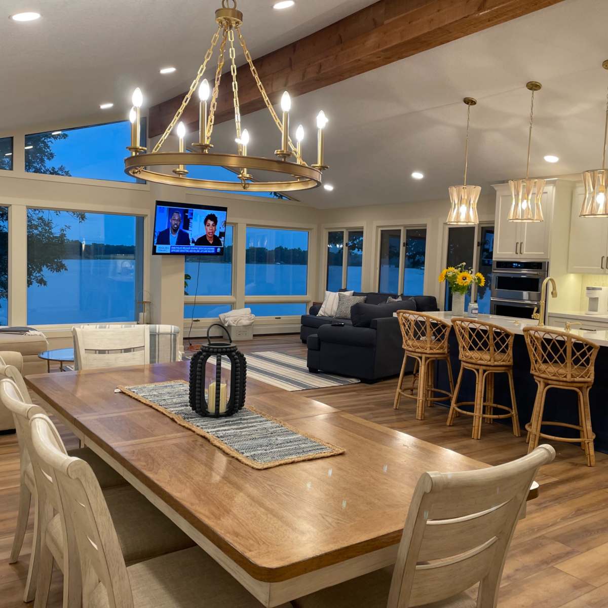 A modern living and dining area with large windows overlooking a body of water, featuring a dining table, comfortable seating, and a television mounted on the wall.