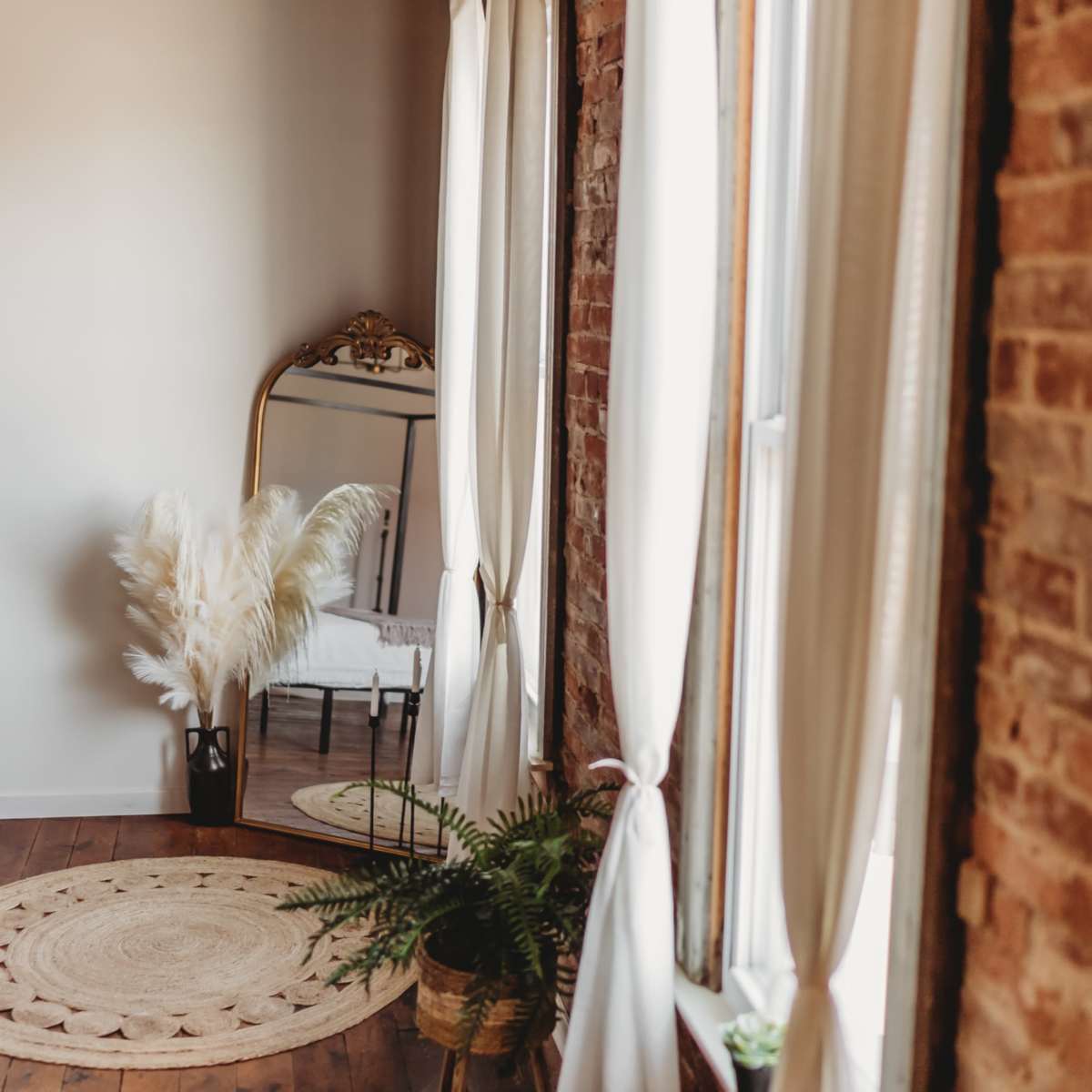 A well-lit room features a large mirror, a rattan rug, potted plants, and floor-to-ceiling windows with sheer curtains.