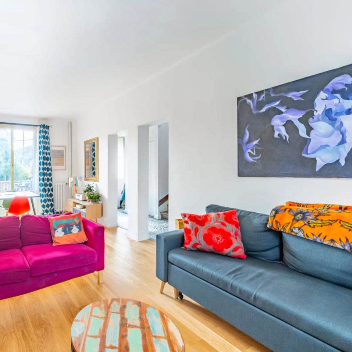 A bright living room features a pink sofa, a dark blue sofa, a round coffee table, and colorful artwork on the walls, with large windows letting in natural light.