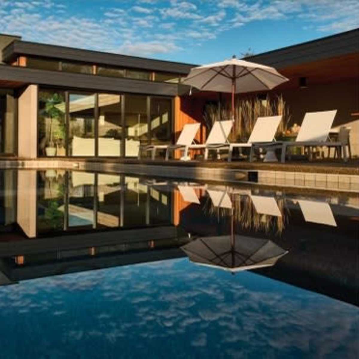A modern house with large windows overlooks a swimming pool surrounded by lounge chairs and an umbrella, reflecting the sky and trees.