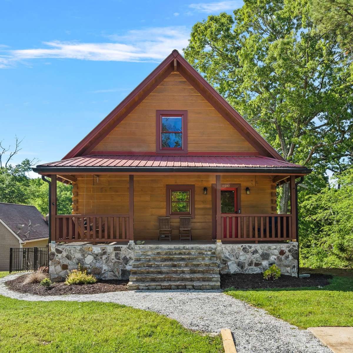 A wooden cabin with a gable roof and stone steps is surrounded by green trees and a grassy yard.