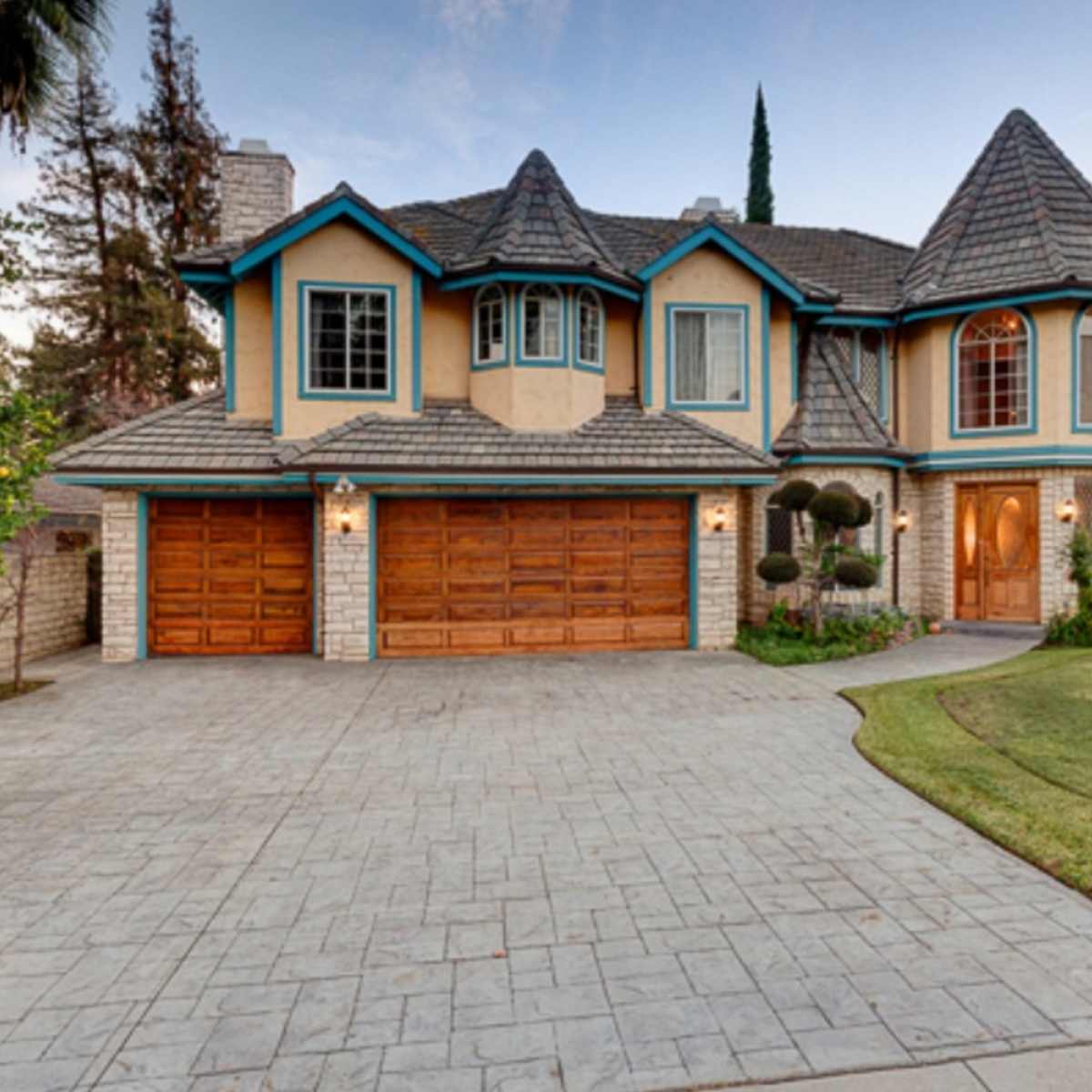 A large, two-story house with a steeply pitched roof, prominent front windows, and a landscaped yard featuring manicured shrubs and a stamped concrete driveway.