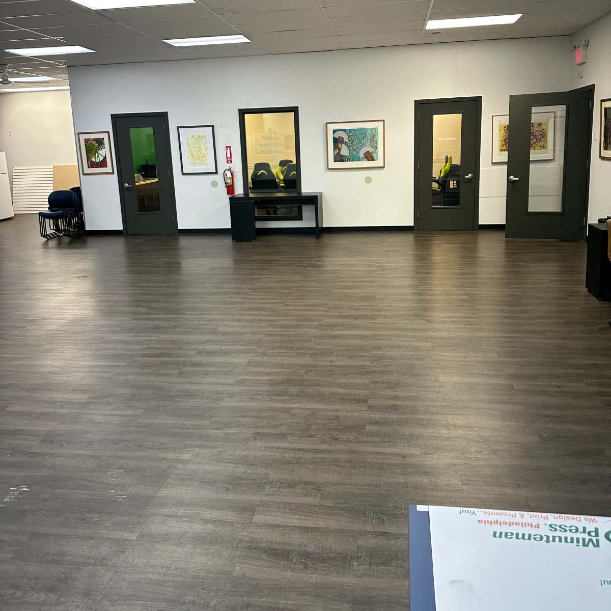 A spacious interior room with wood flooring, several doors, framed artworks on the walls, and a few chairs visible in the background.