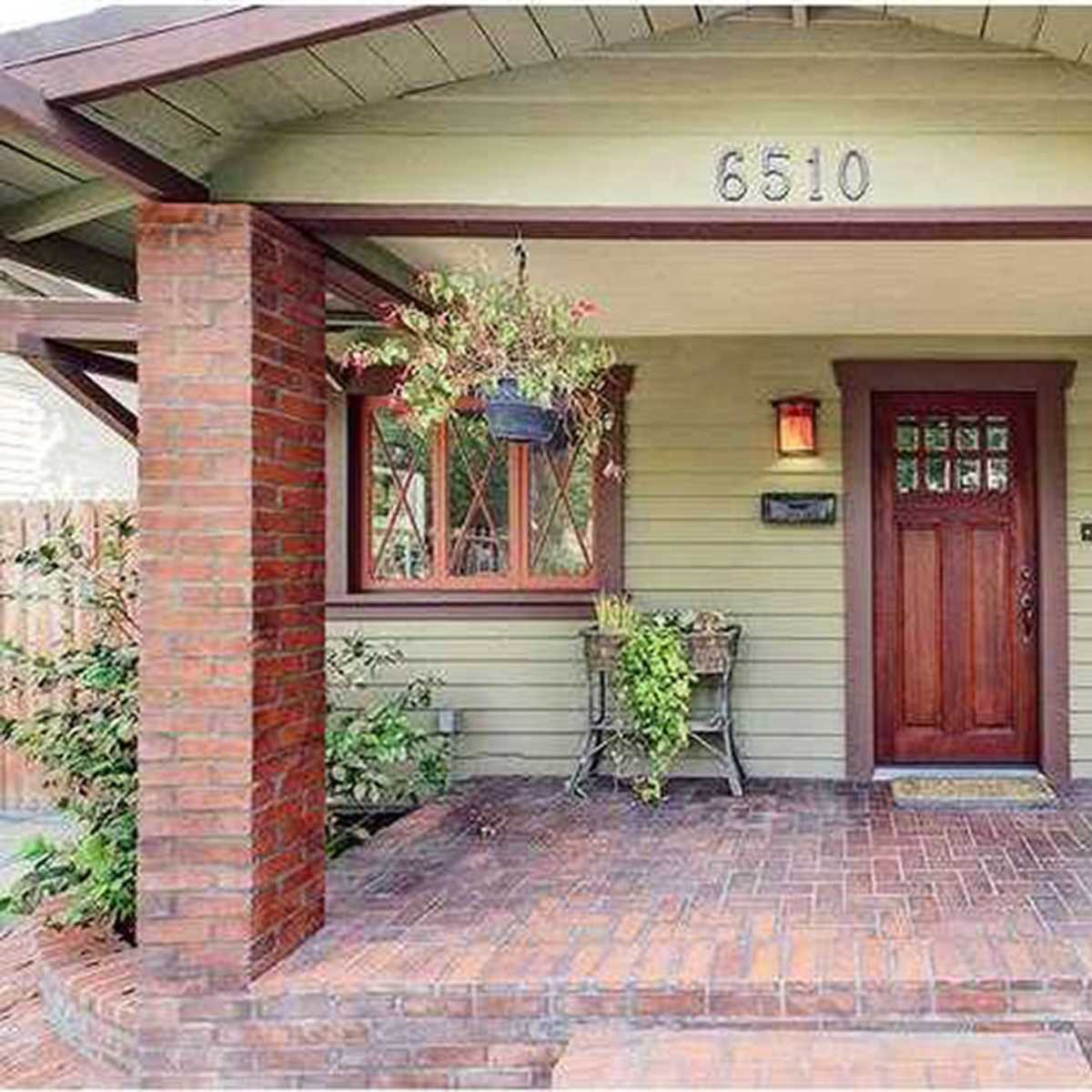 A front porch of a green bungalow with red brick pillars and a wooden door, featuring potted plants and a welcome mat.