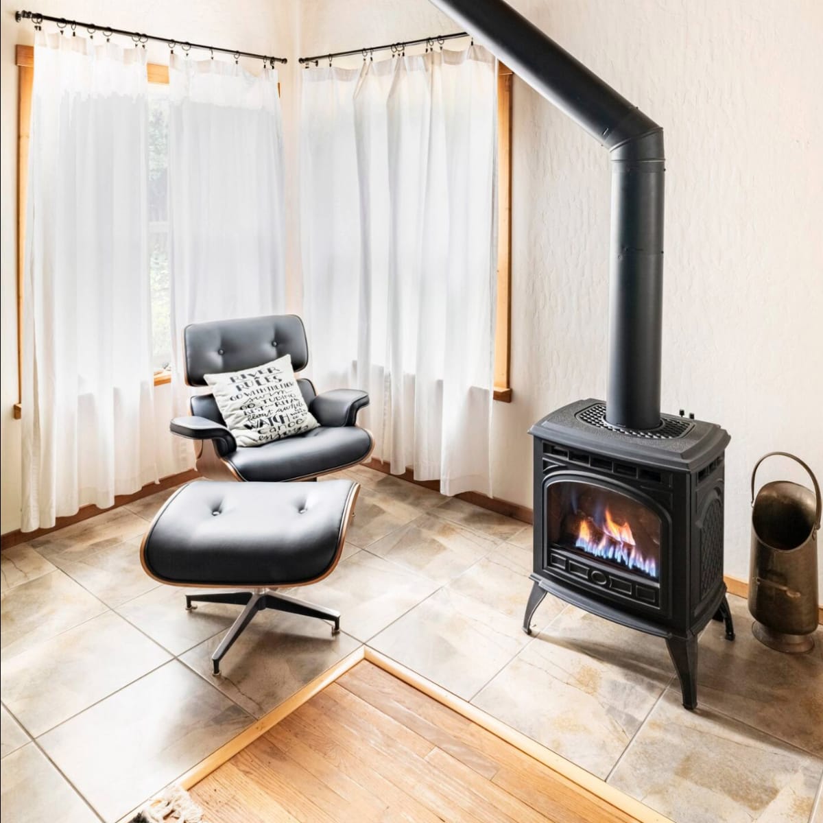 A modern black wood stove is positioned in a bright room with tall windows and a stylish armchair nearby.