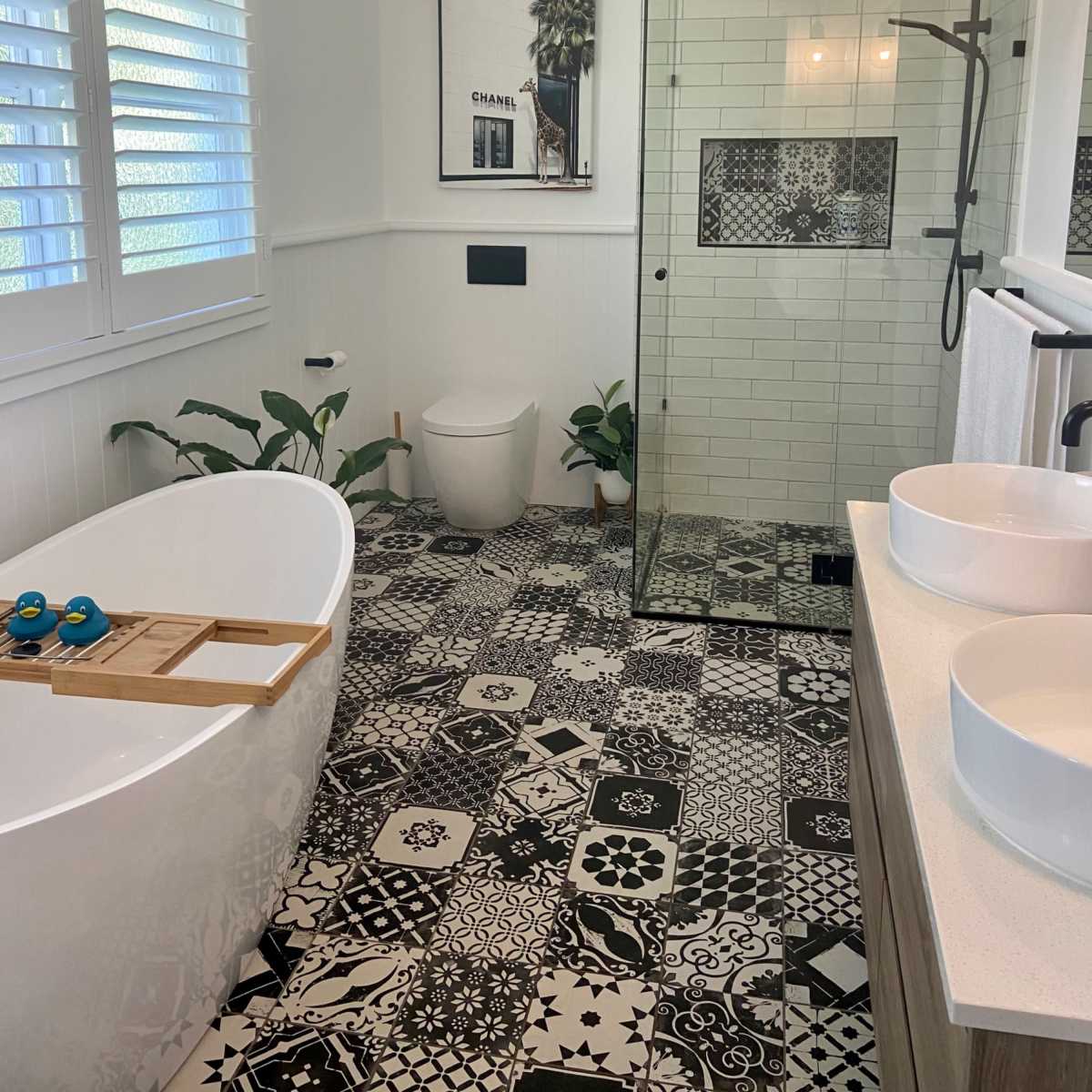 A modern bathroom features a freestanding bathtub, a walk-in shower with glass walls, patterned tile flooring, and greenery accents.