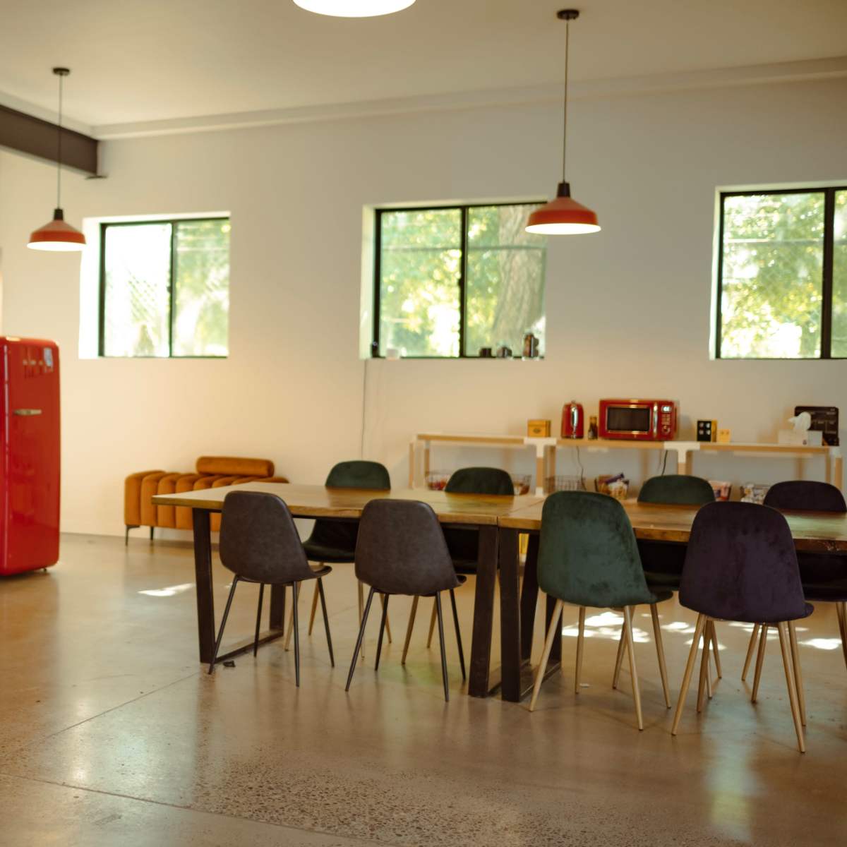 A modern kitchen and dining area featuring a red refrigerator, a wooden dining table with upholstered chairs, and large windows allowing natural light.