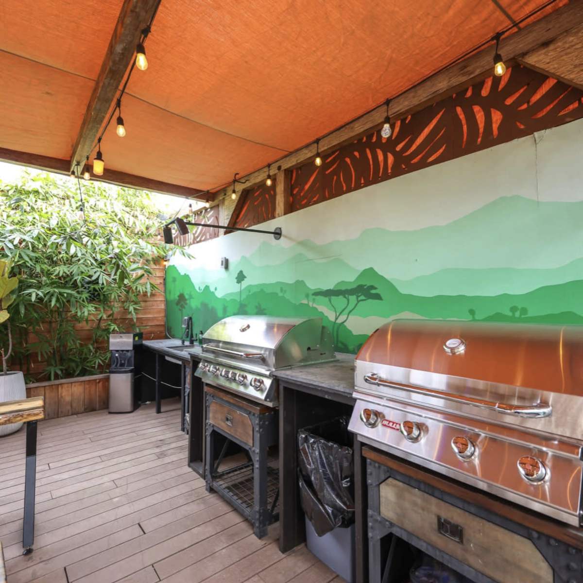 An outdoor grilling area featuring several stainless steel grills under an orange canopy, with a mural of green hills on the wall.