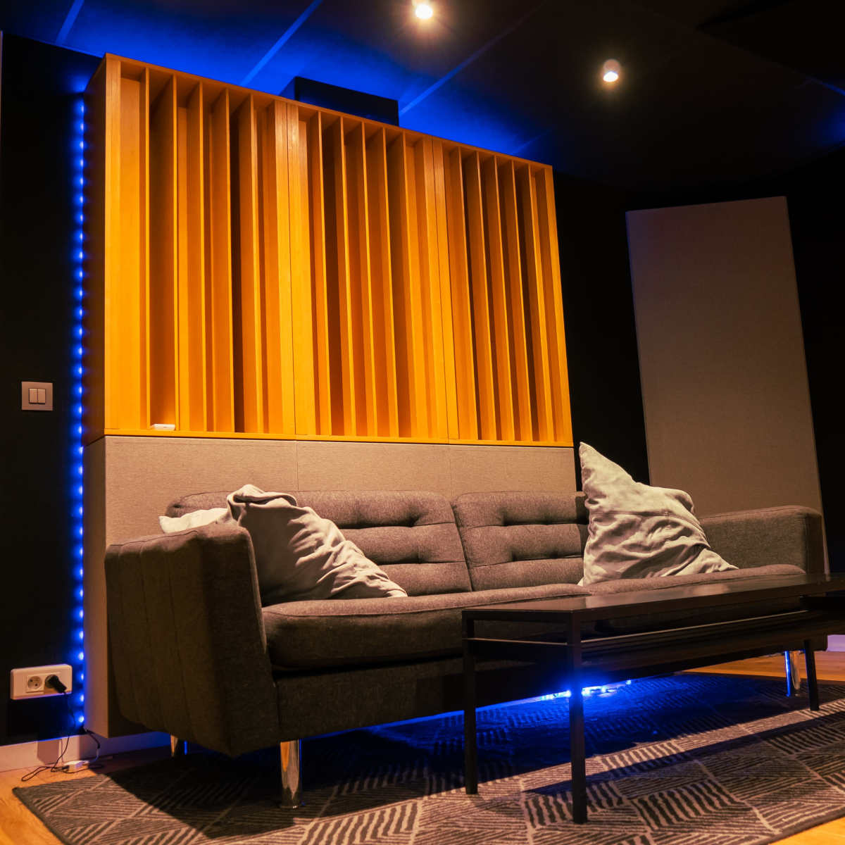 A modern sound studio with a gray sofa, a black coffee table, and wooden acoustic panels on the walls.