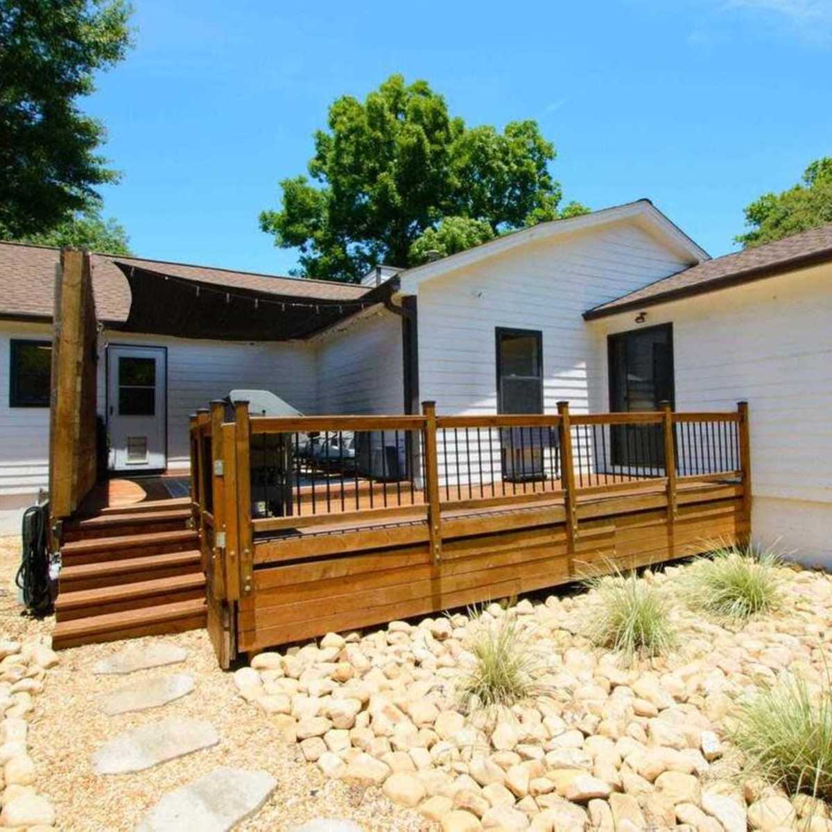 A single-story house with a wooden deck, surrounded by a landscaped area featuring gravel and ornamental plants.