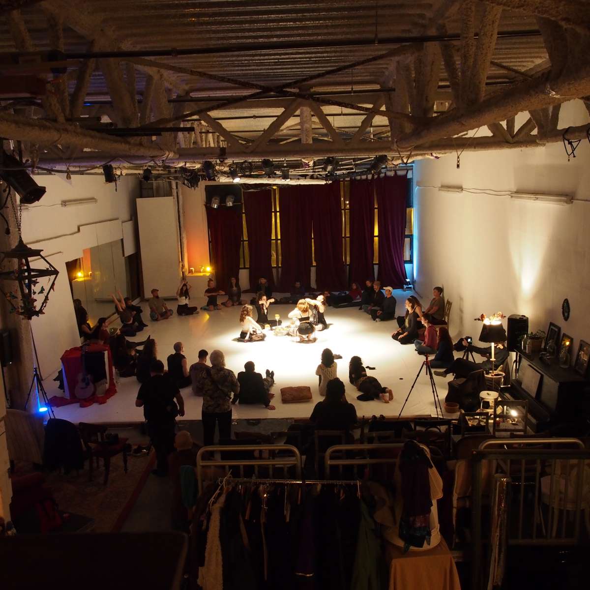 A large group of people is seated around a performance space in a dimly lit room with raised ceilings and red curtains.