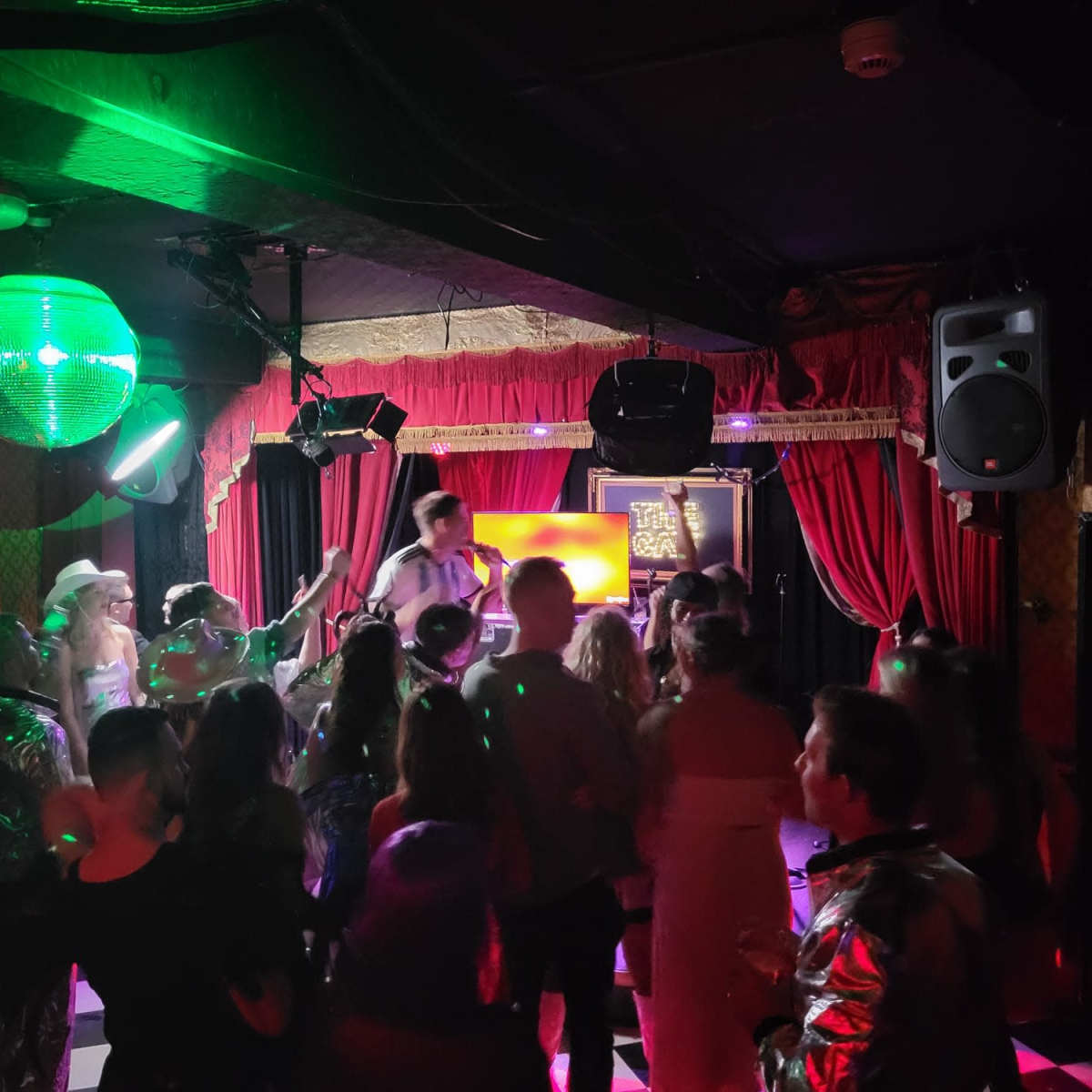 A lively crowd dances under a disco ball in a dimly lit bar with colorful lights.