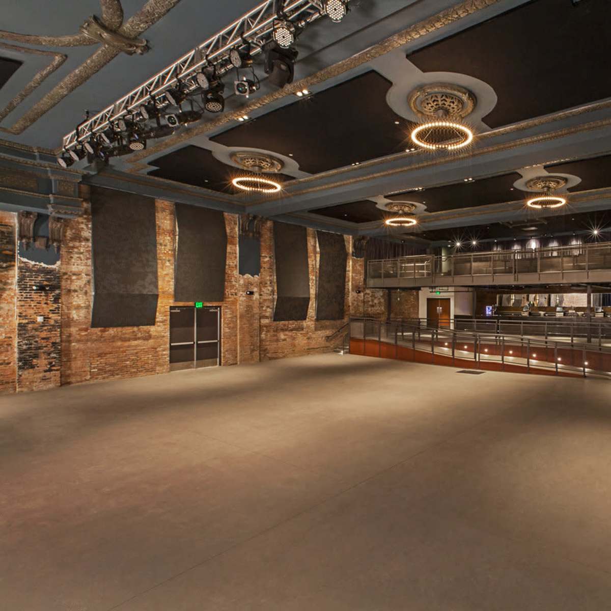 A large, empty concert venue featuring a stage with overhead lighting and a balcony area, with exposed brick walls and a polished floor.