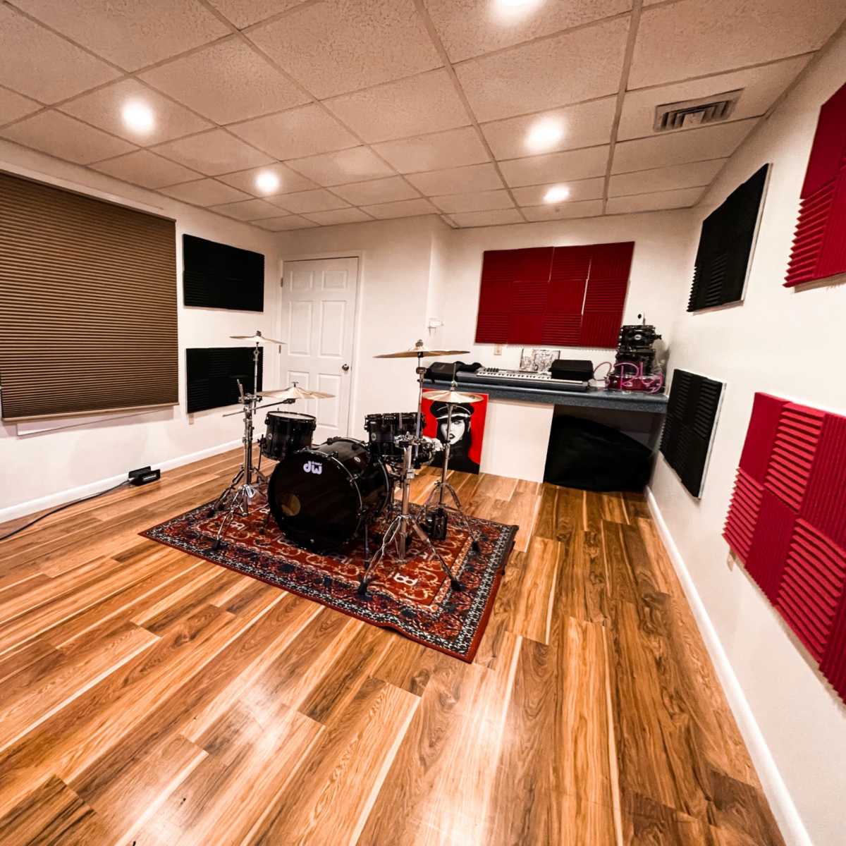 A music rehearsal room with a black drum set on a rug, surrounded by soundproofing panels on the walls and wooden flooring.