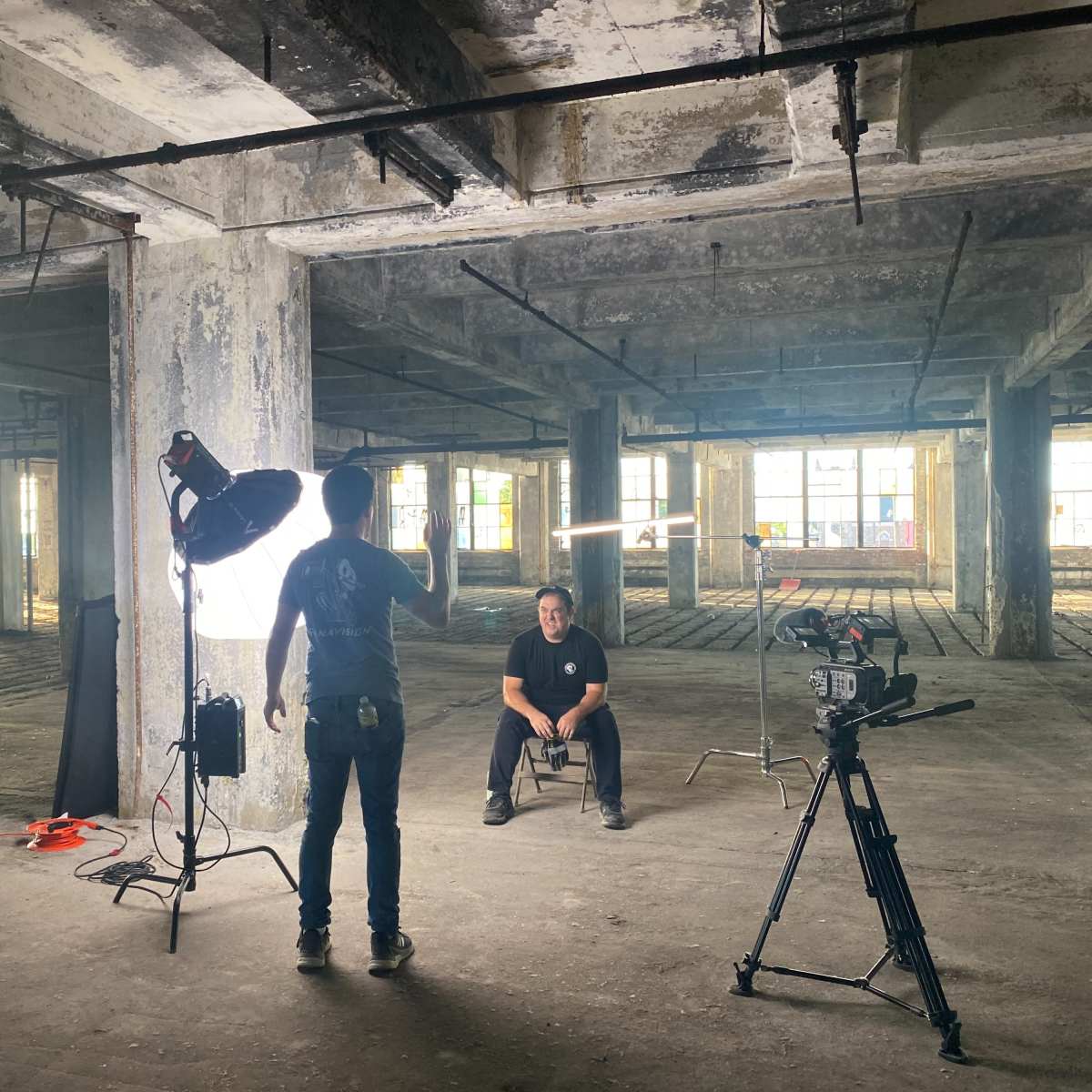 A person sits on a chair in a dilapidated industrial space while another individual operates a camera and lighting equipment.