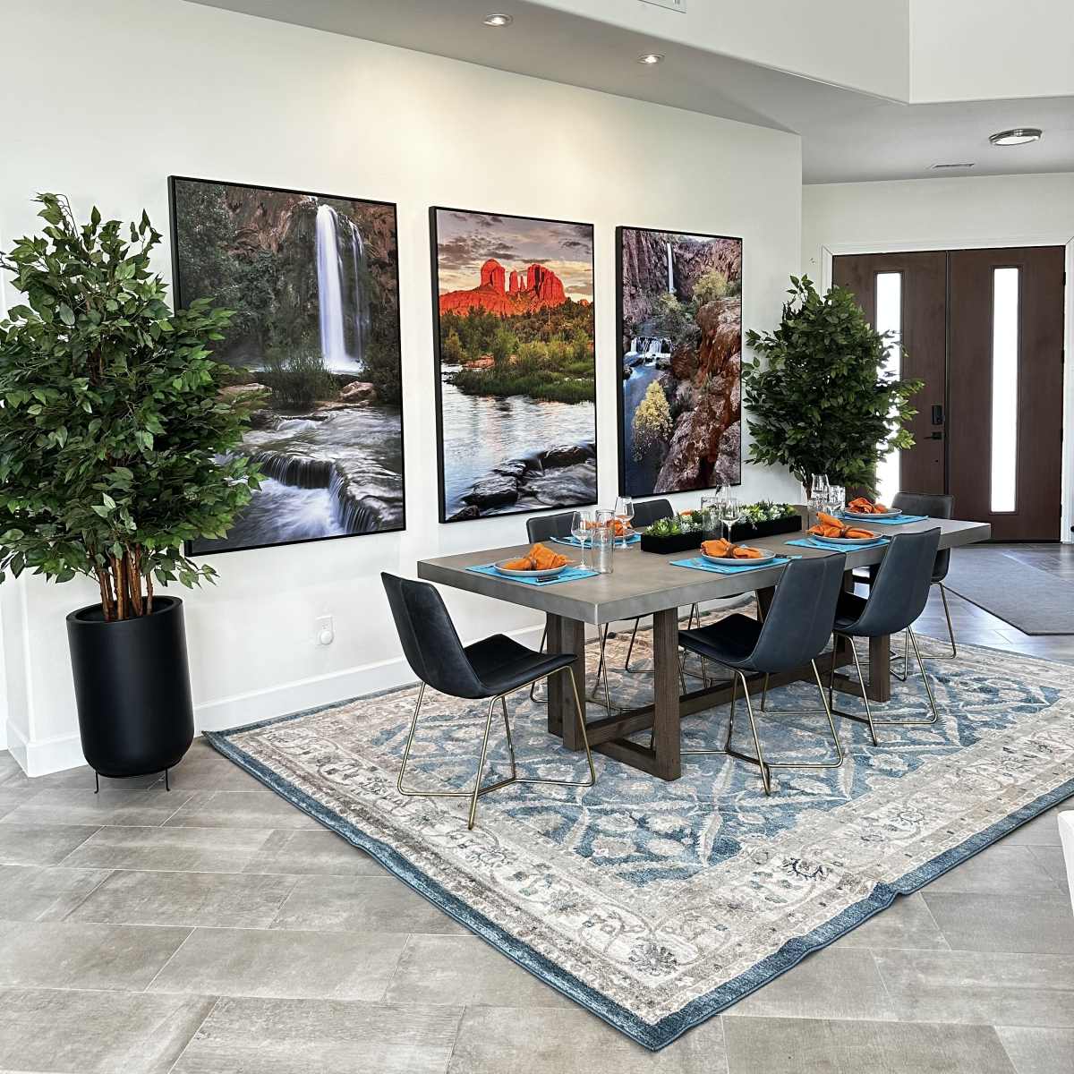 A modern dining area features a table set for four, flanked by two green plants, with large landscape photographs on the wall and a decorative rug beneath.
