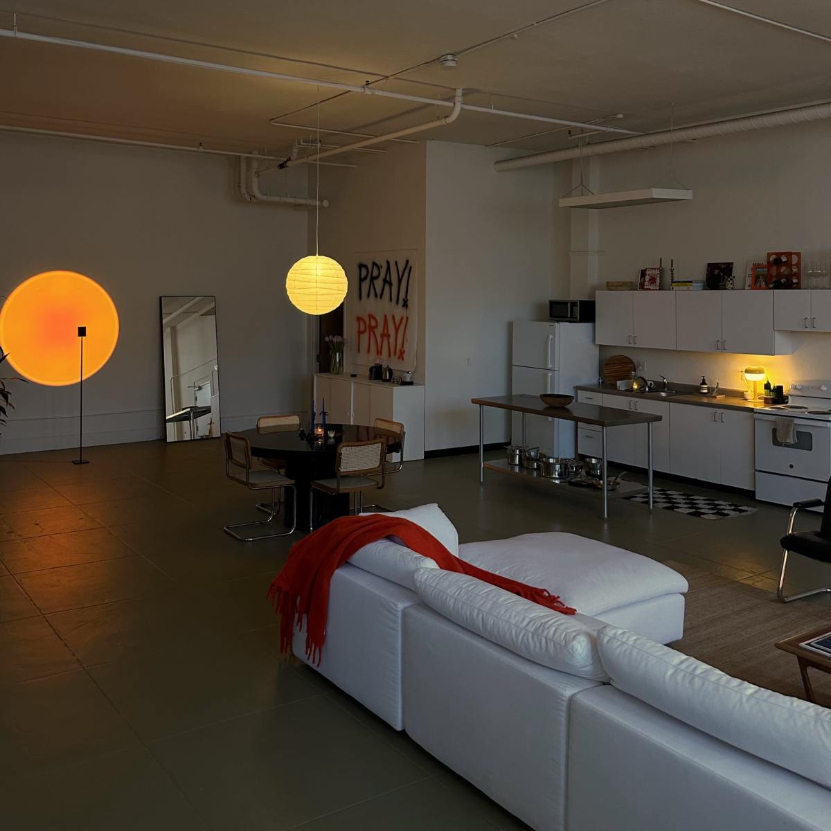 A spacious, modern apartment with a white couch, a black and white checkered rug, and a kitchen area illuminated by warm light, featuring an abstract circular wall decoration and a large mirror.