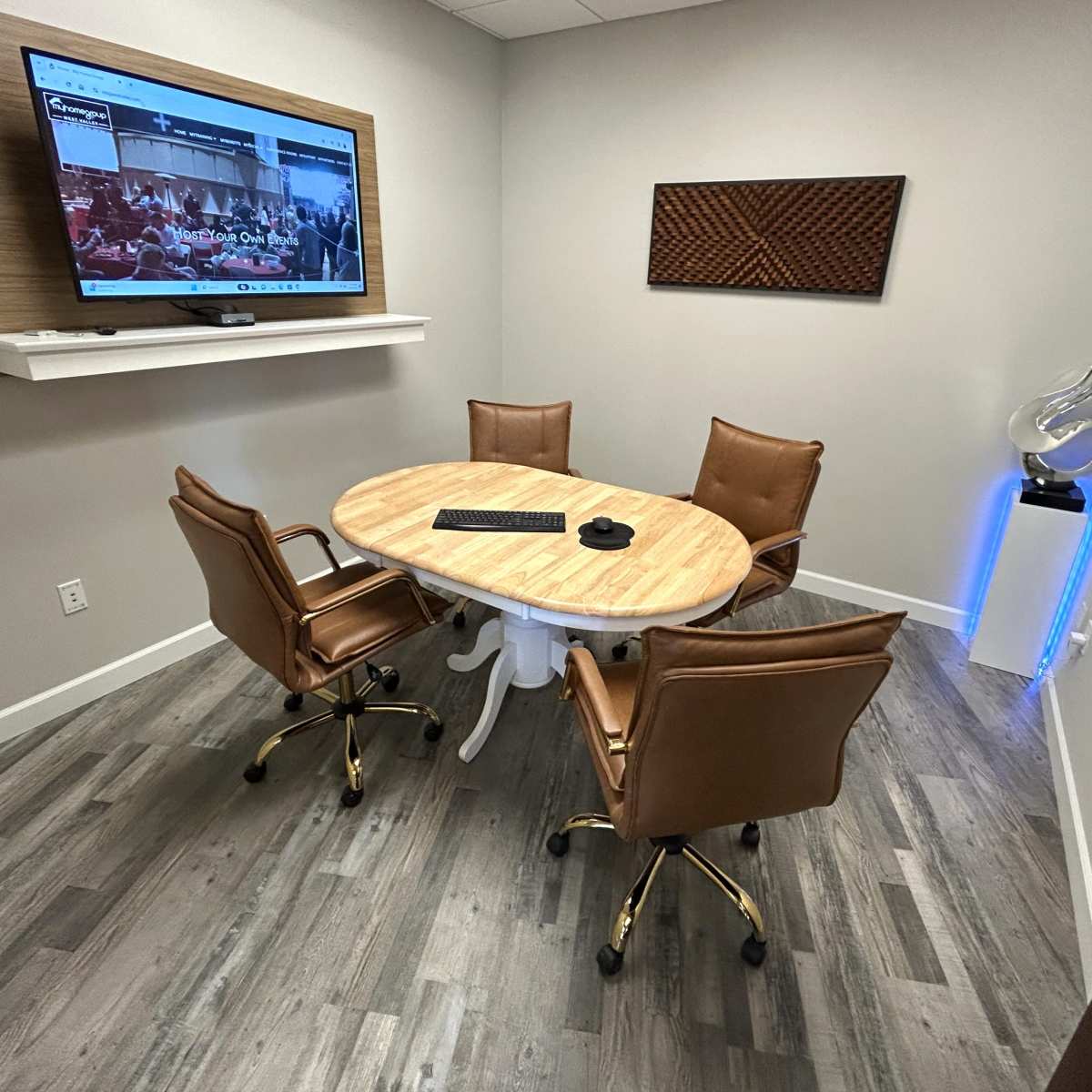 A small conference room with a circular wooden table surrounded by four brown leather chairs, a wall-mounted television, and modern decorative elements.