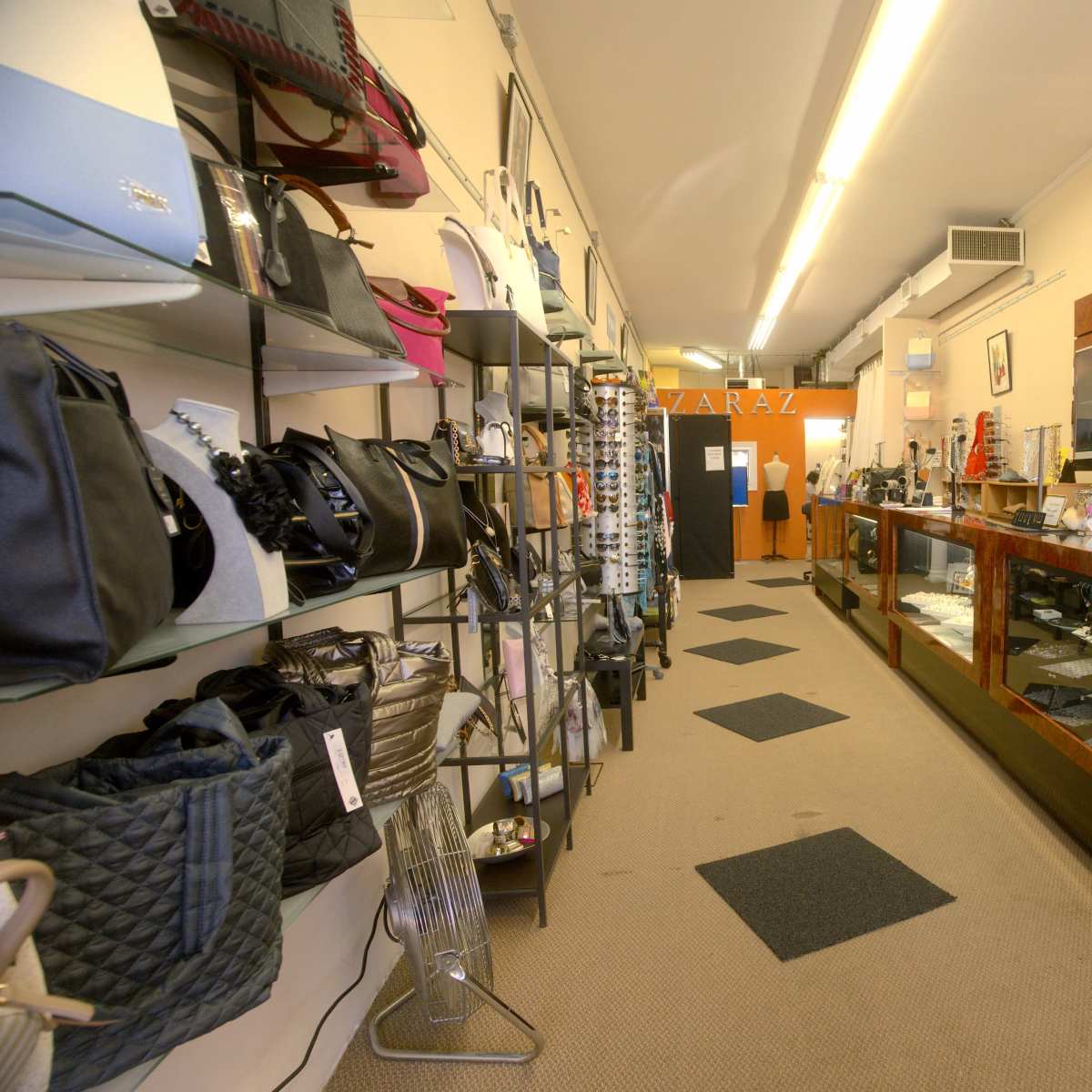 A narrow retail store interior with displays of handbags and jewelry along both sides of the aisle.