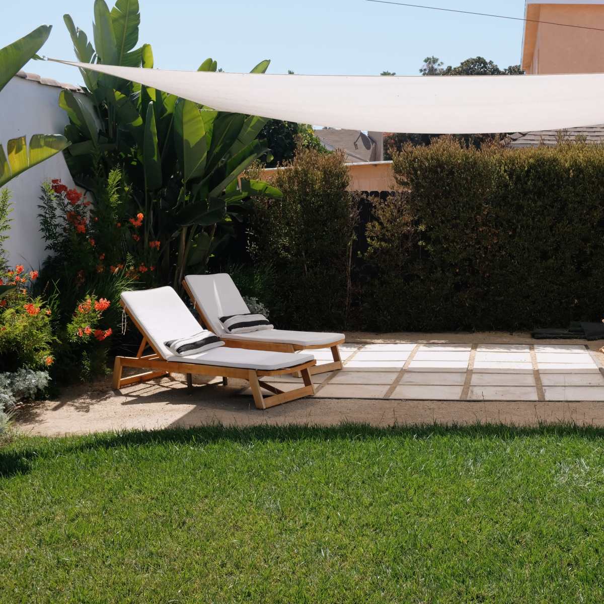 A landscaped backyard with two lounge chairs and a seating area under a fabric sunshade, surrounded by greenery and flowering plants.