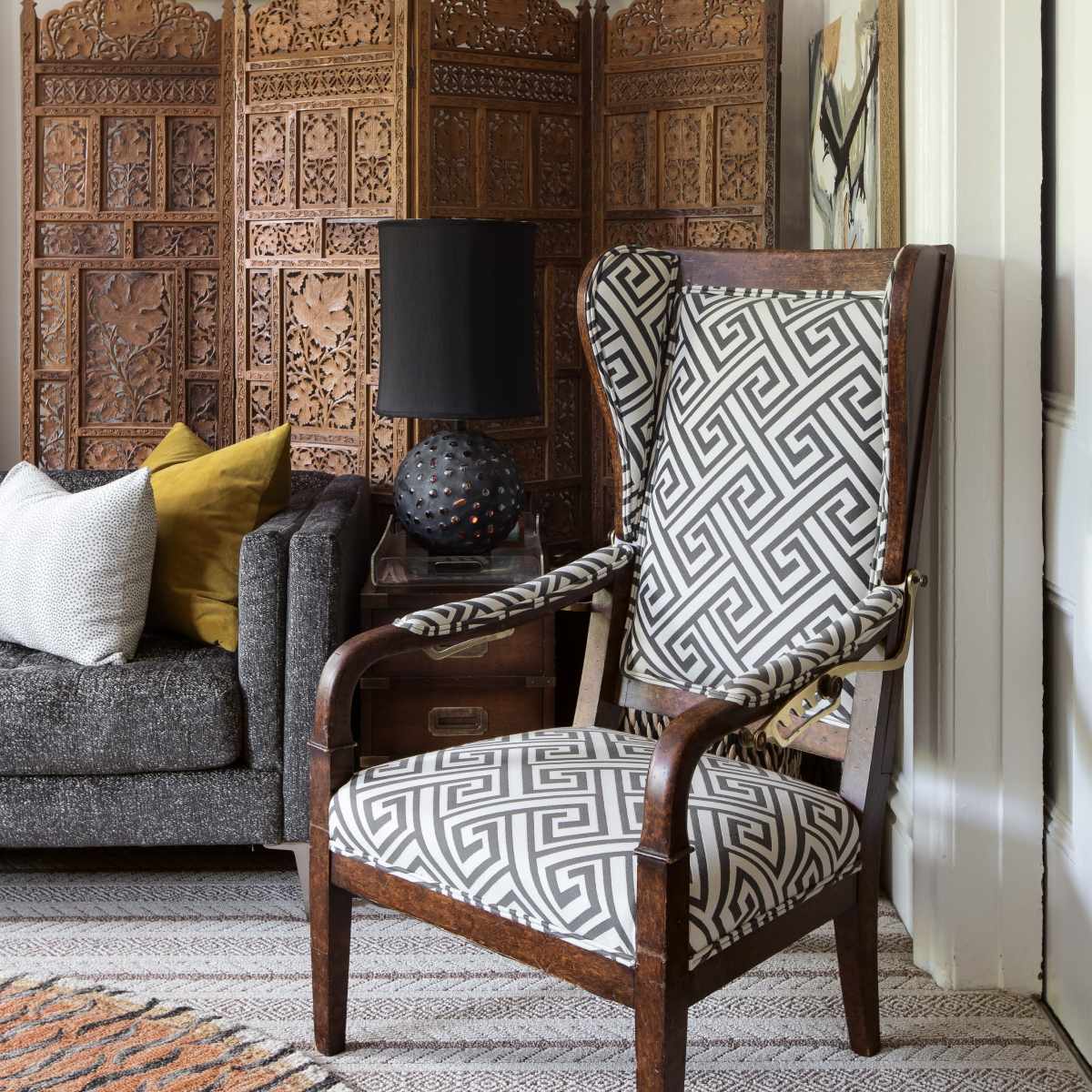 The scene features a patterned armchair beside a textured gray sofa, with wooden decorative panels and abstract artwork in the background.