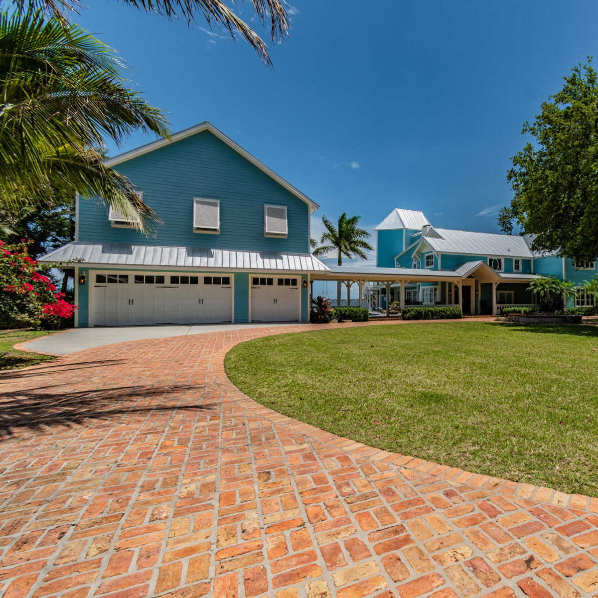 A large blue house with multiple gables, surrounded by a well-kept lawn and palm trees, with a brick pathway leading to the front entrance.