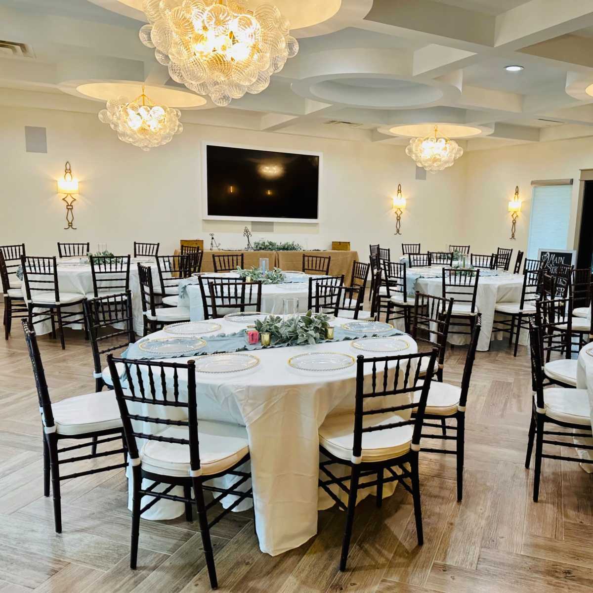 An elegantly arranged dining area with several round tables covered in white tablecloths, surrounded by black chairs, and decorated with greenery and light fixtures.