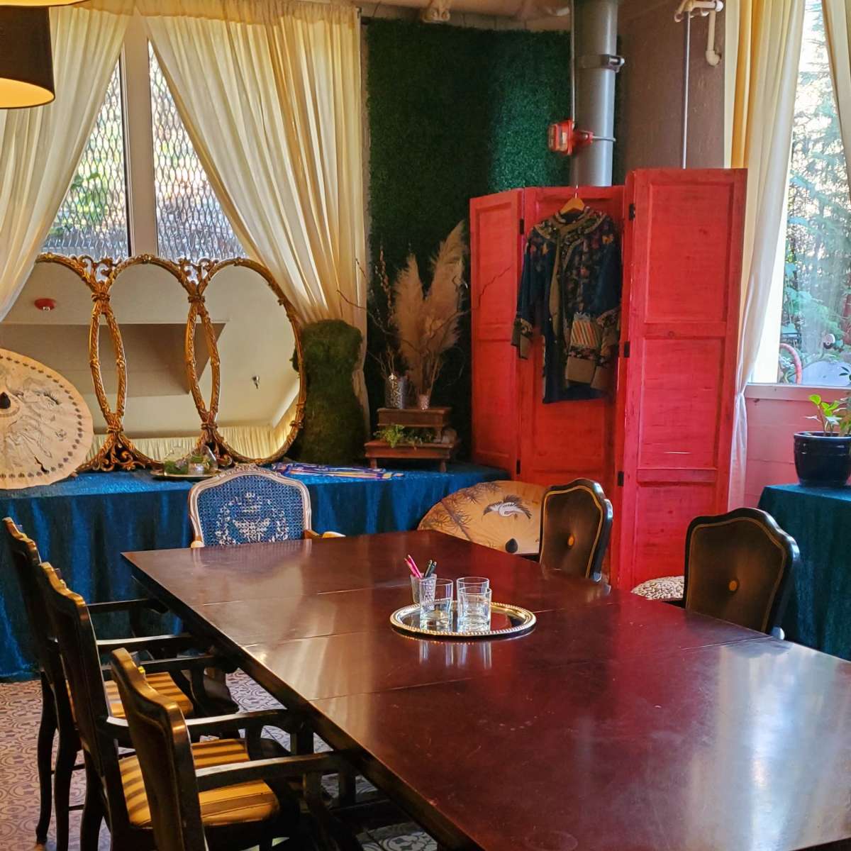 A long wooden dining table with multiple chairs is set in a room featuring a decorative red cabinet, mirrors, and greenery.