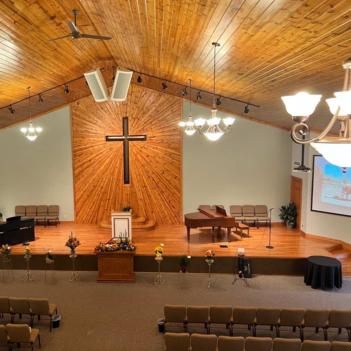 An interior view of a church featuring a wooden cross on the back wall, rows of chairs, and a stage with a piano and floral arrangements.