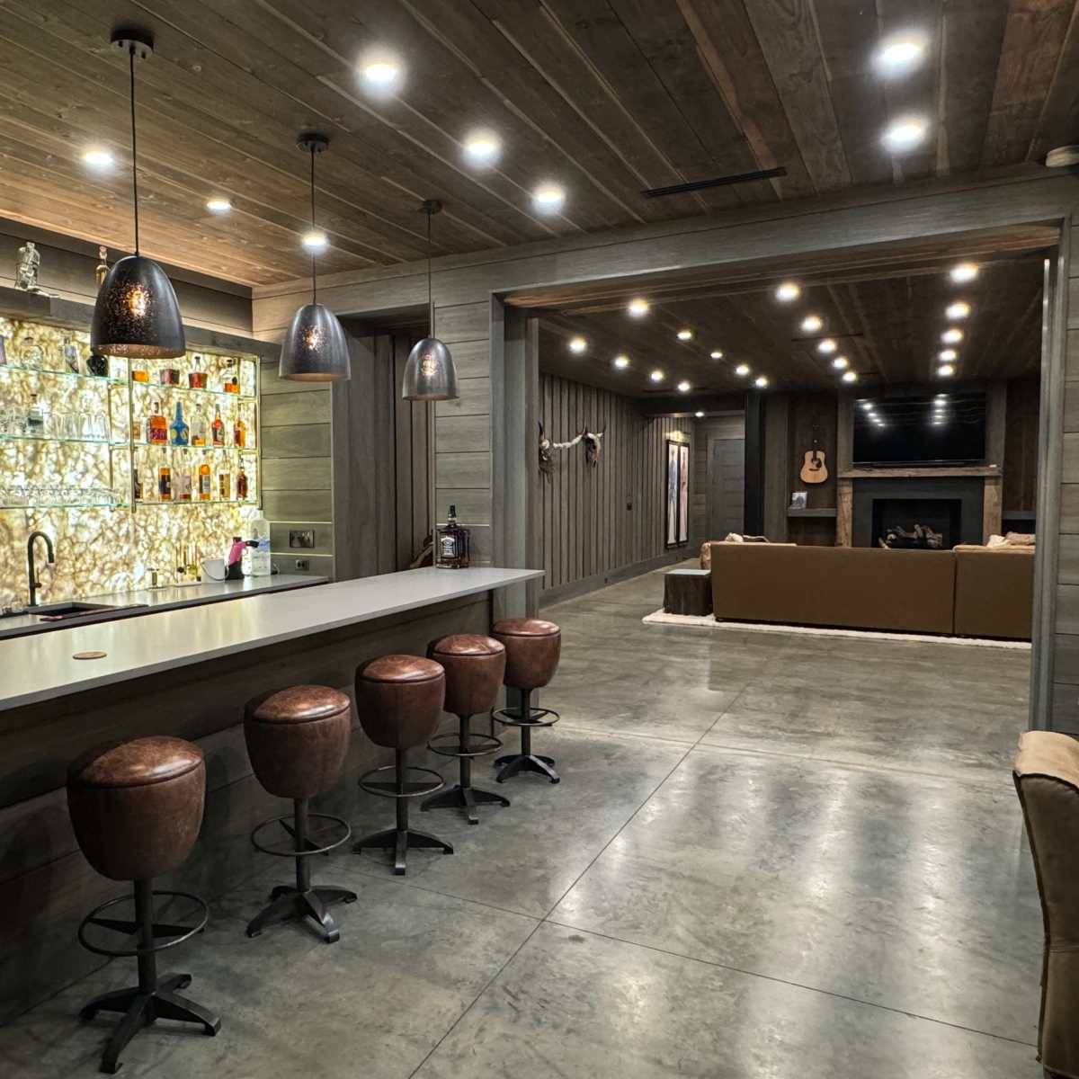 A modern interior space featuring a bar with tall stools, a well-lit backlit shelf displaying various bottles, and a living area with a large sofa and a television.