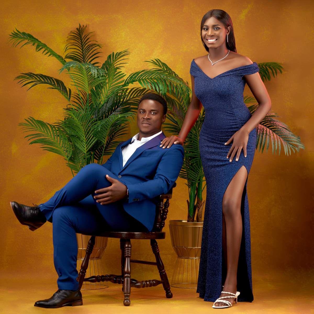 A man in a blue suit sits on a wooden chair while a woman in a sparkling blue gown stands next to him against a backdrop of a golden wall and leafy plants.