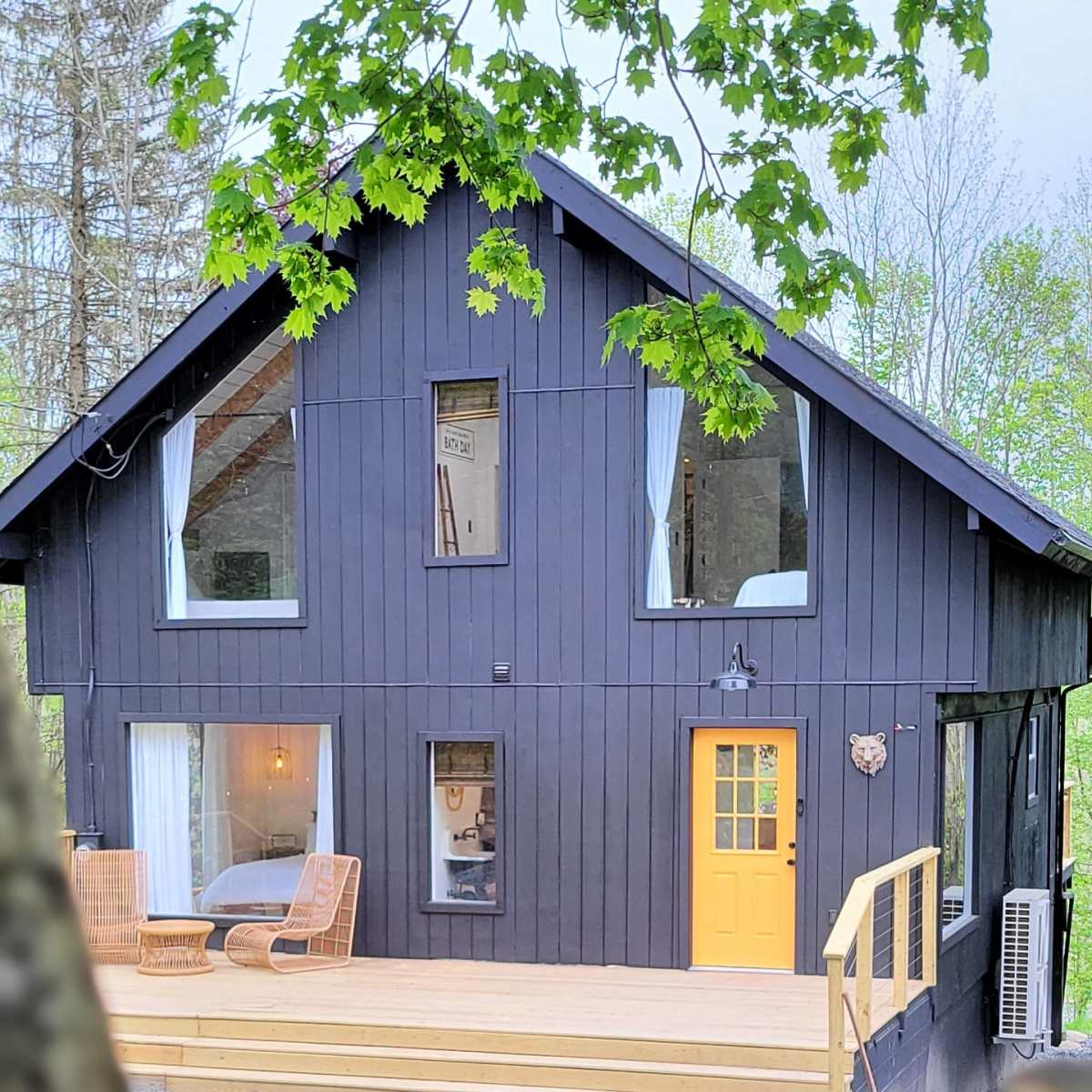 A black wooden house with a yellow front door and large windows is set against a backdrop of trees.