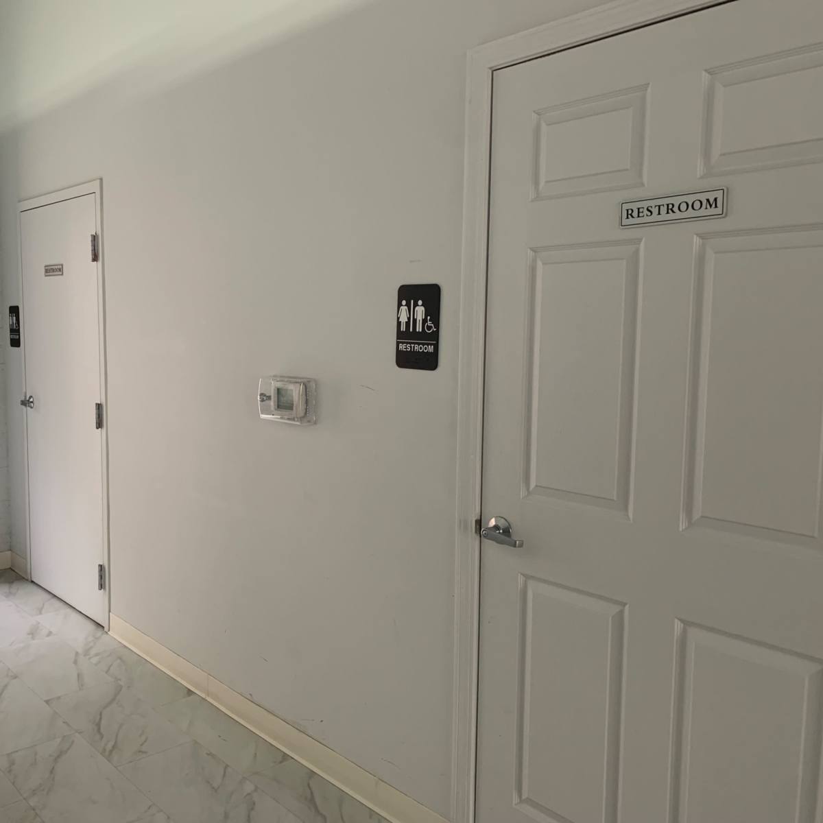 A clean, white corridor with two doors labeled for restrooms, one on the right and another on the left, and an exit door at the far end.