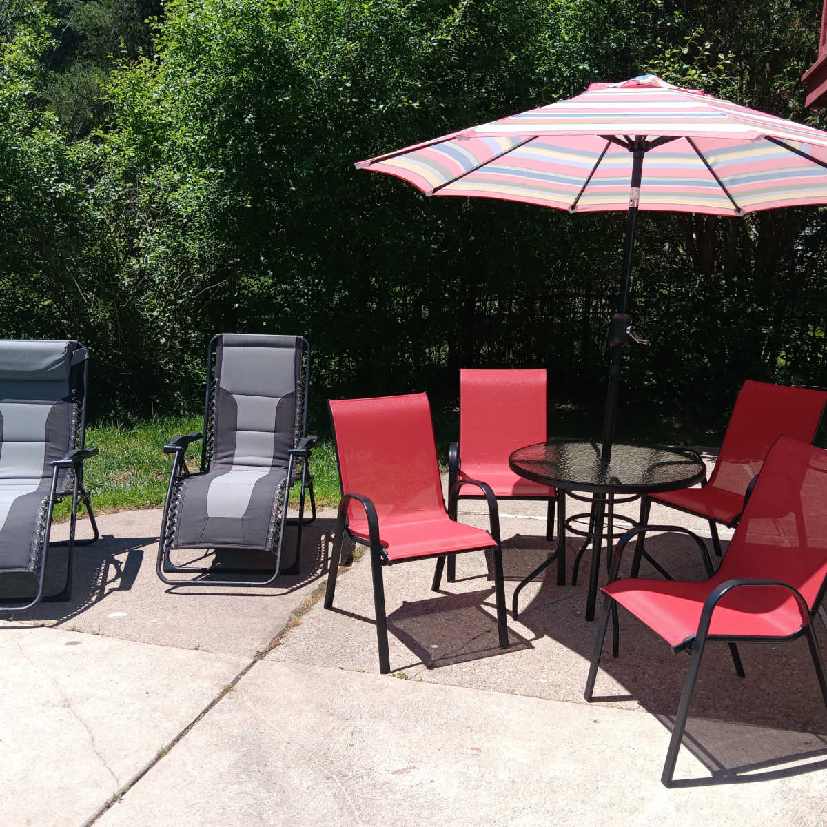 A patio area with two reclining chairs, four red dining chairs, a glass table, and a striped umbrella, surrounded by greenery.