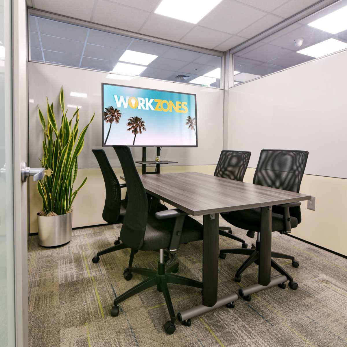 A conference room features a wooden table, four black swivel chairs, a large screen displaying "WORKZONES," and a tall plant in a pot.