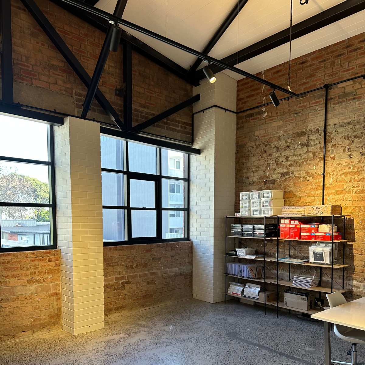 An industrial-style room with exposed brick walls, large windows, and a metal shelf filled with boxes and papers in one corner.
