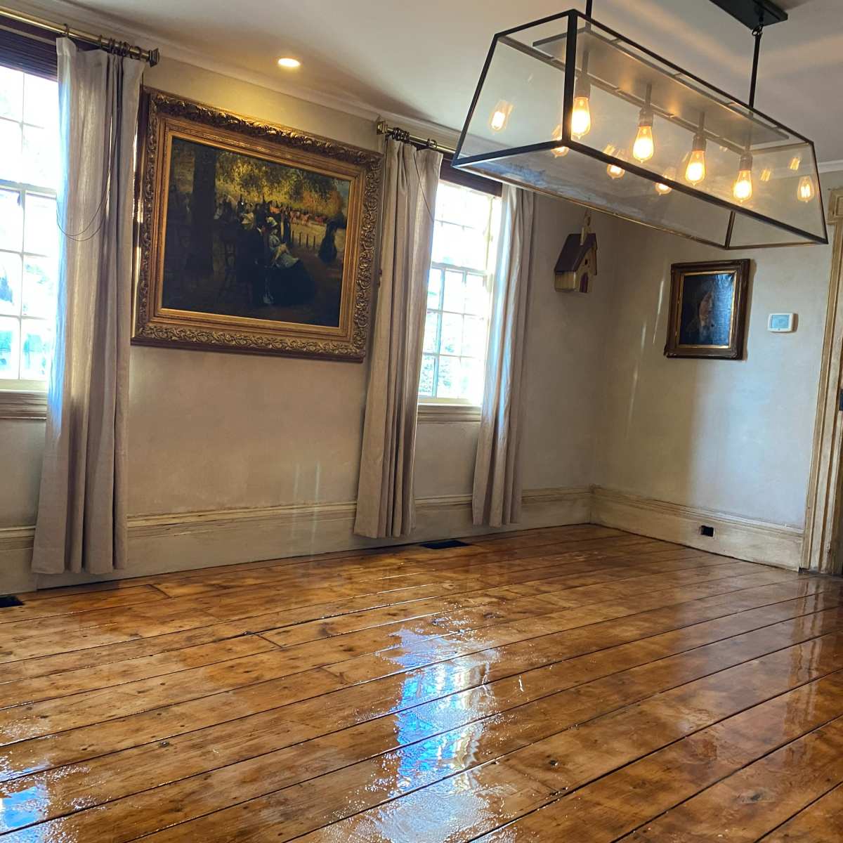 The room features a polished wooden floor, large windows, and a modern light fixture hanging from the ceiling, complemented by classical artwork on the walls.