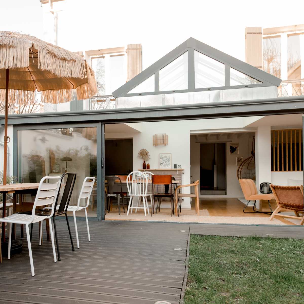 A modern patio features a large thatched umbrella, a wooden dining table with chairs, and a glass wall that opens to an interior space.