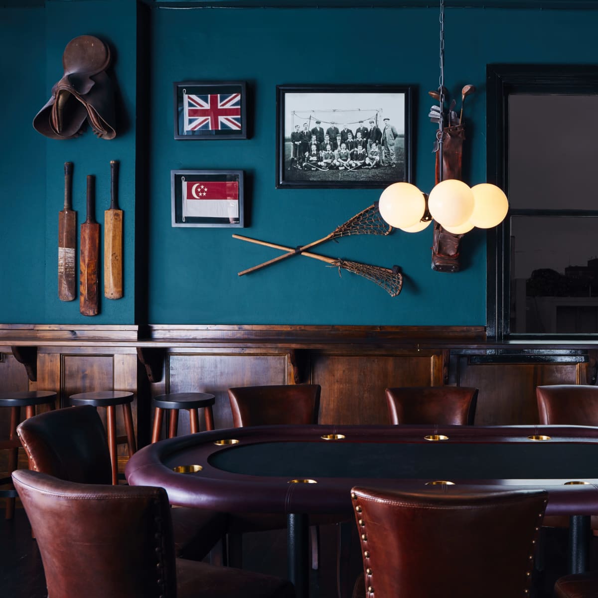 A dimly lit room featuring a round poker table surrounded by leather chairs, with framed sports memorabilia and a large screen on the walls.