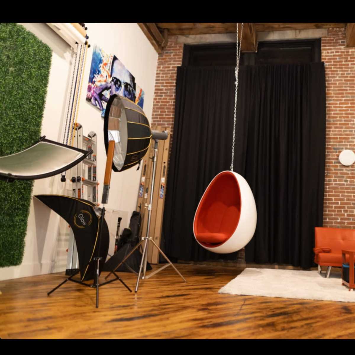 A modern living space features a hanging white egg chair, a red leather sofa, a wooden coffee table, and a backdrop of exposed brick walls.