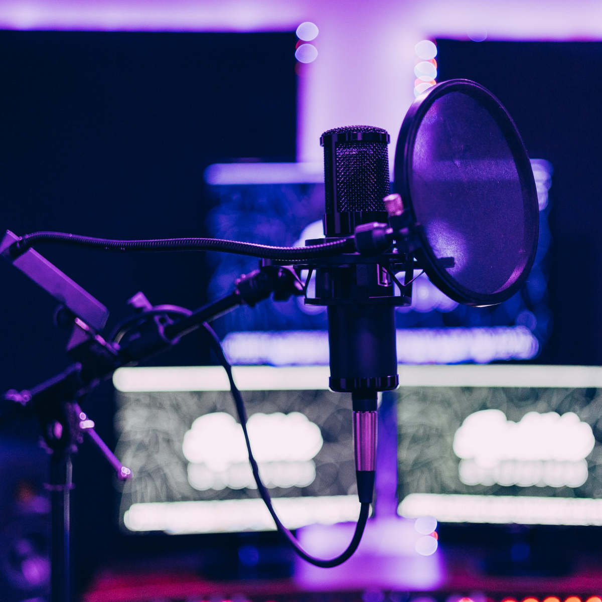 A microphone on a stand is positioned in front of two computer screens in a sound studio.