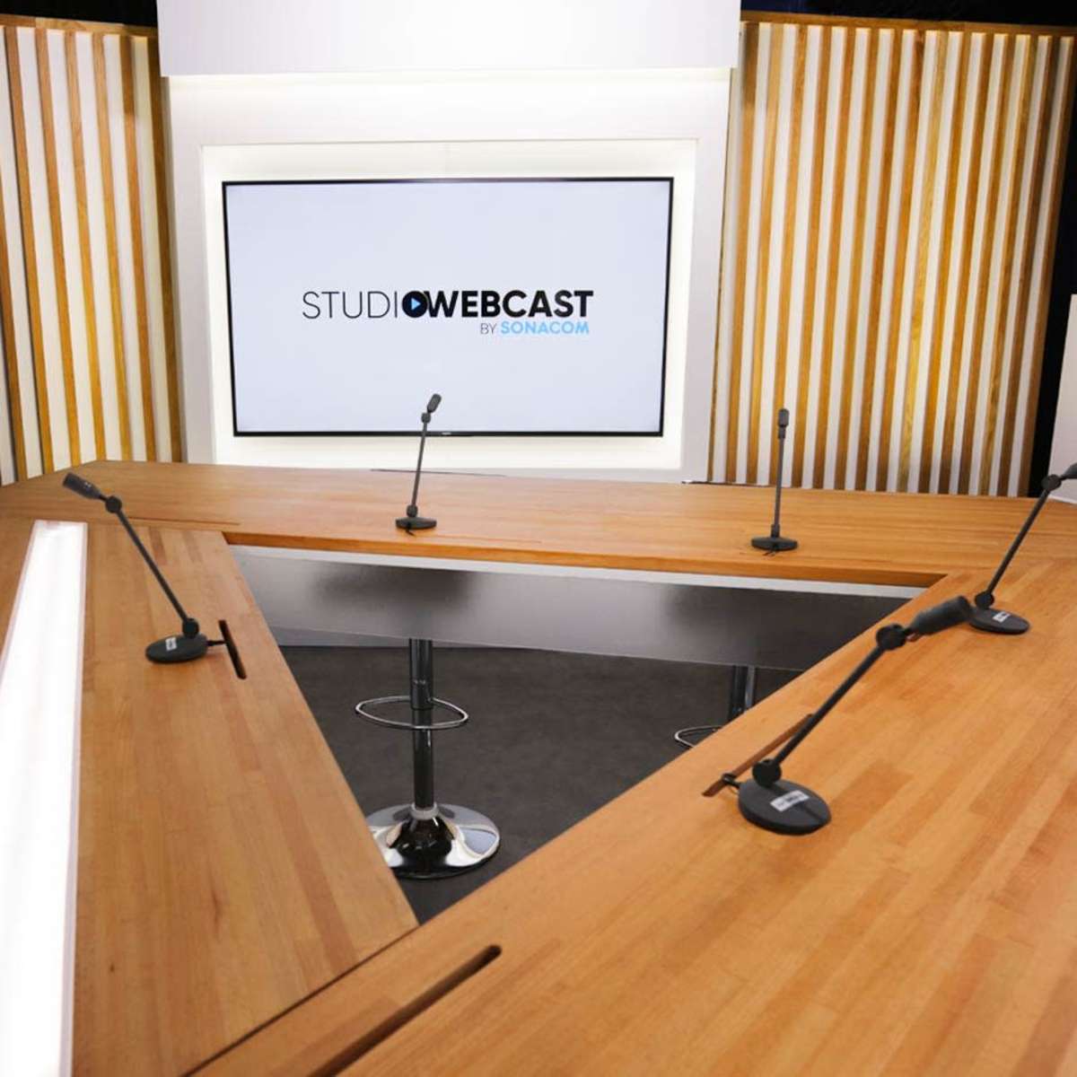 A modern broadcast studio with a large wooden table, multiple microphones, and a screen displaying the logo "STUDIO WEBCAST."