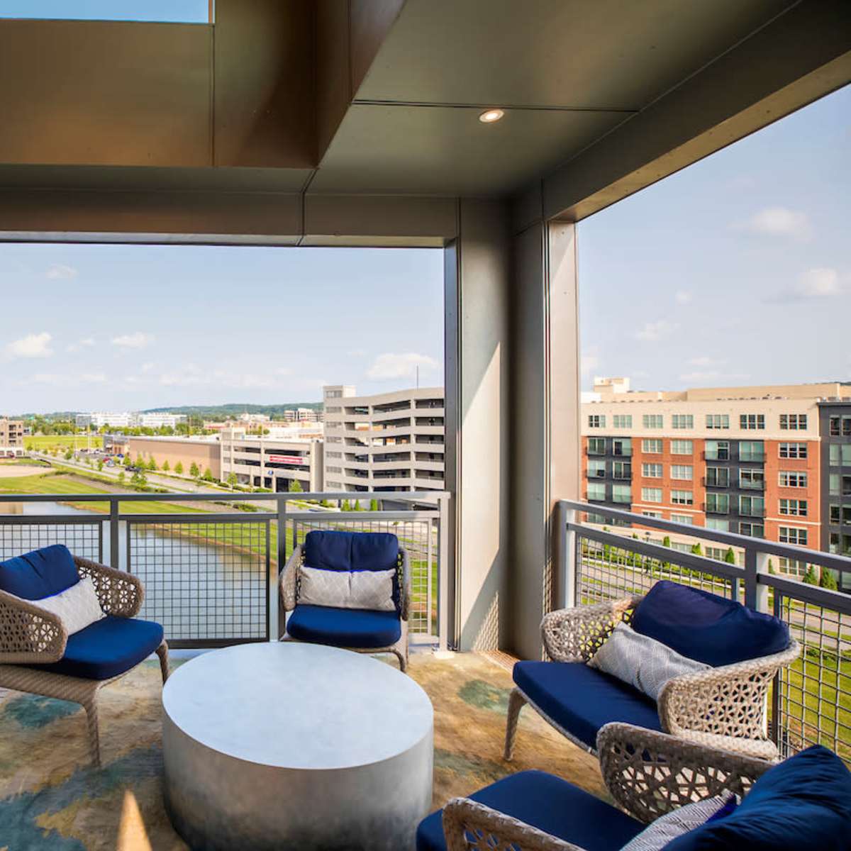 A balcony with modern seating overlooks a green area and parking structures beside a body of water.