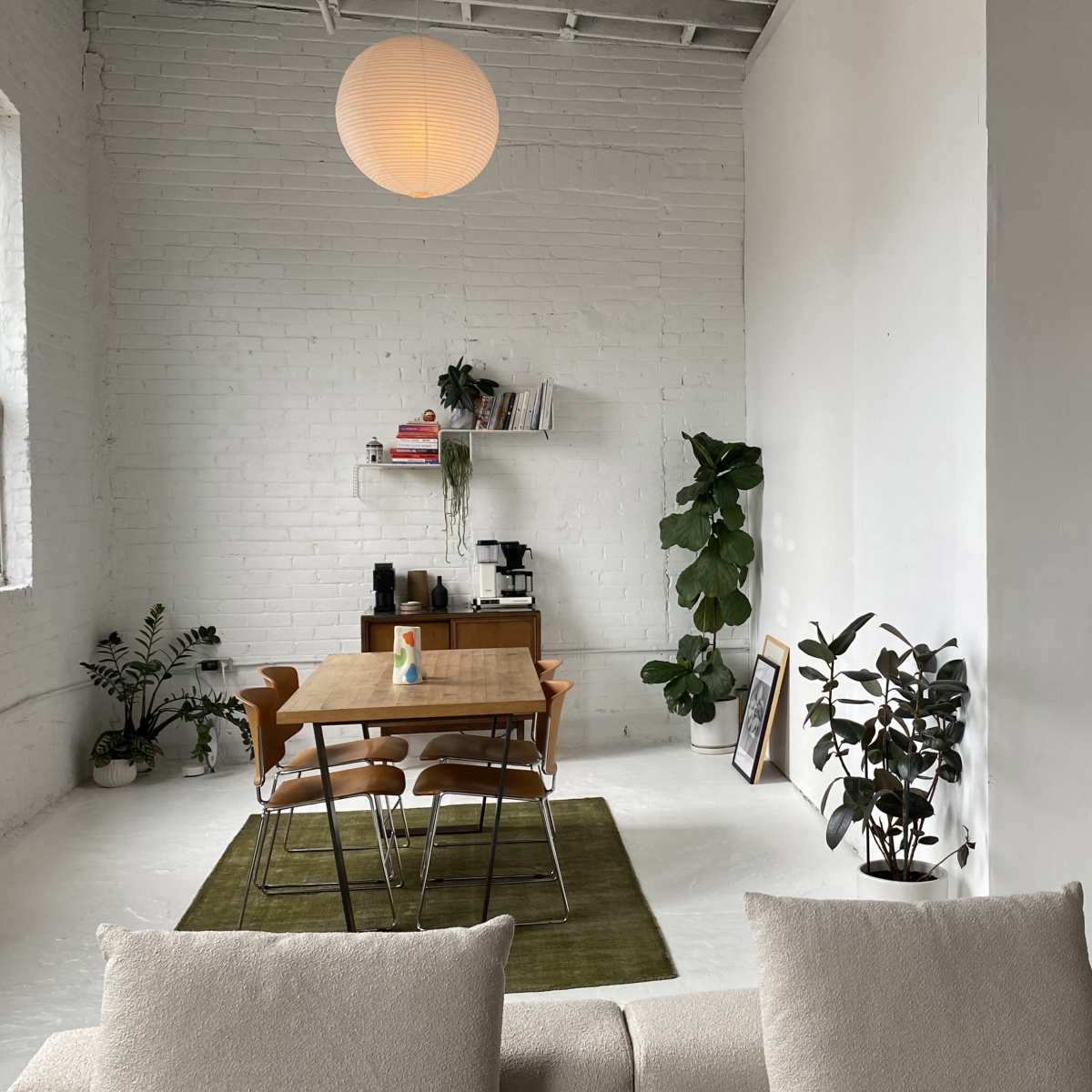 A bright, minimalist room featuring a dining table with chairs, a wall shelf with books and plants, and a sofa in the foreground.