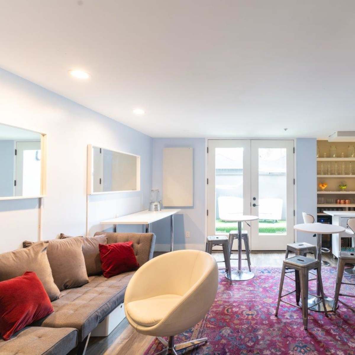 A modern lounge area features a gray sofa, white chairs, small tables, and a bar with glass shelves, leading to a doorway with access to an outdoor space.