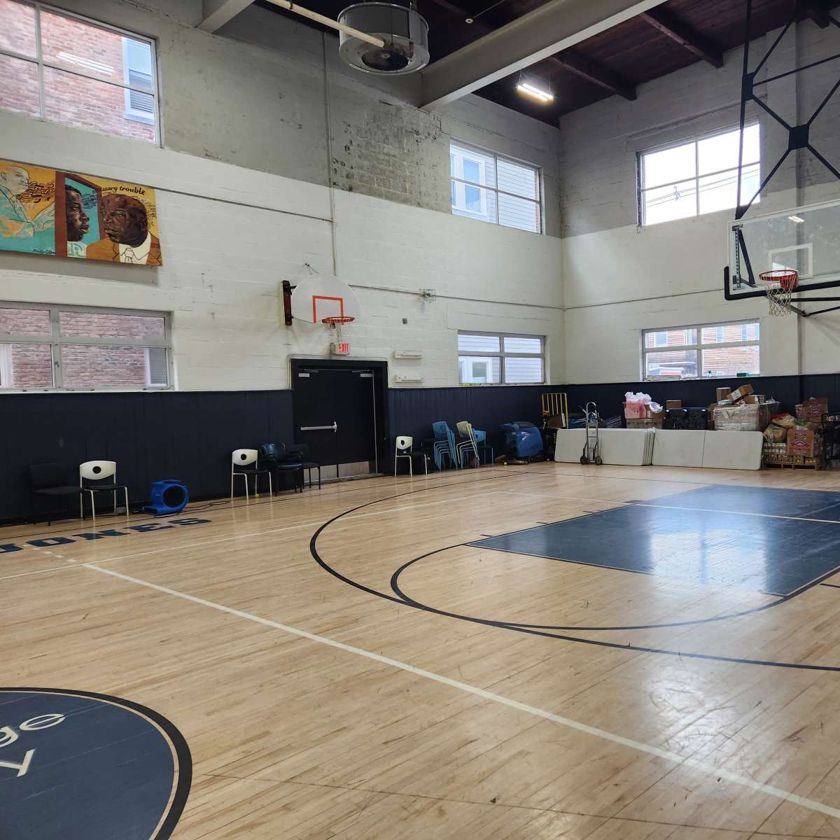 An indoor basketball court with a hoop and some seating along one side, along with boxes and supplies stacked in the background.