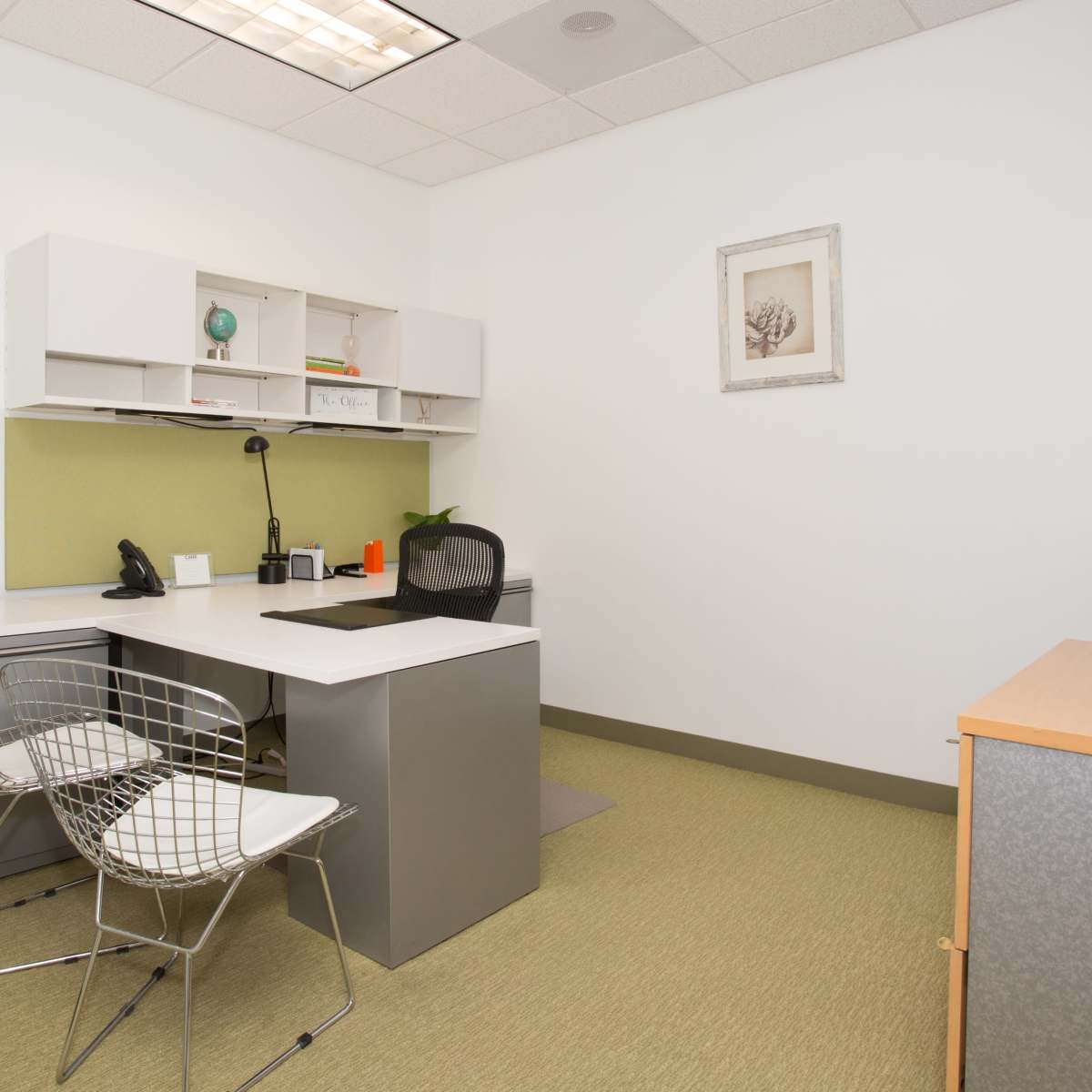 A minimalist office space featuring a desk with a lamp, two wire chairs, a filing cabinet, and a small decorative plant.