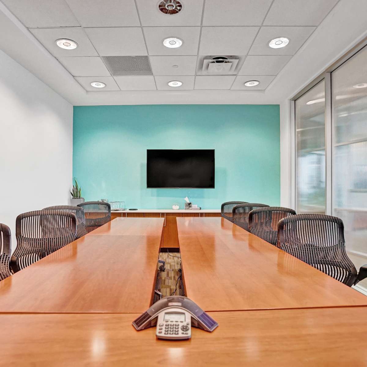A conference room features a long wooden table surrounded by black mesh chairs, with a television mounted on a teal wall and glass partitions.