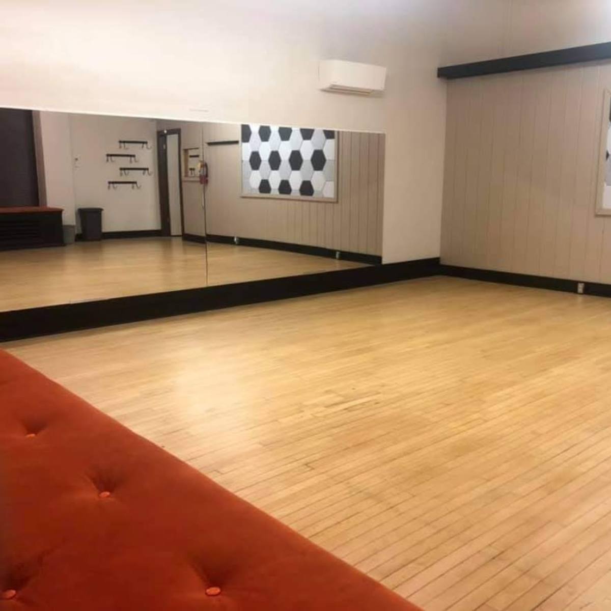 A spacious dance studio with a wooden floor, large mirrors along one wall, and a wall featuring a hexagonal black and white design.