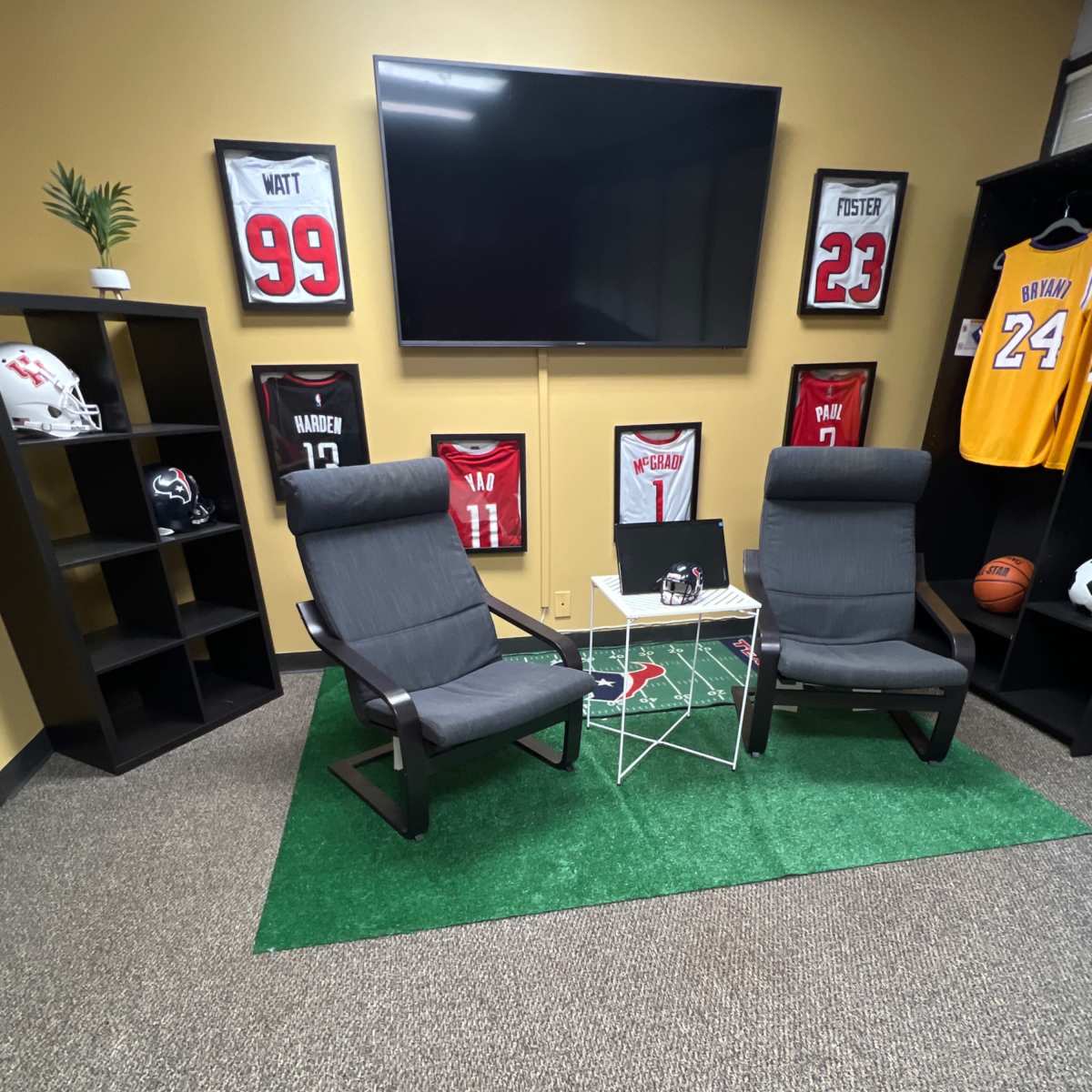 The scene features two gray chairs facing a small table on a green carpet, surrounded by framed jerseys on the walls and a large TV mounted above.